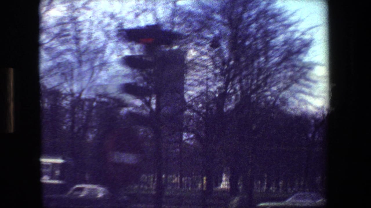 Paris street scene from a moving 8mm camera, showing a red traffic light, winter trees, cars, and a tall building partly obscured by blur.