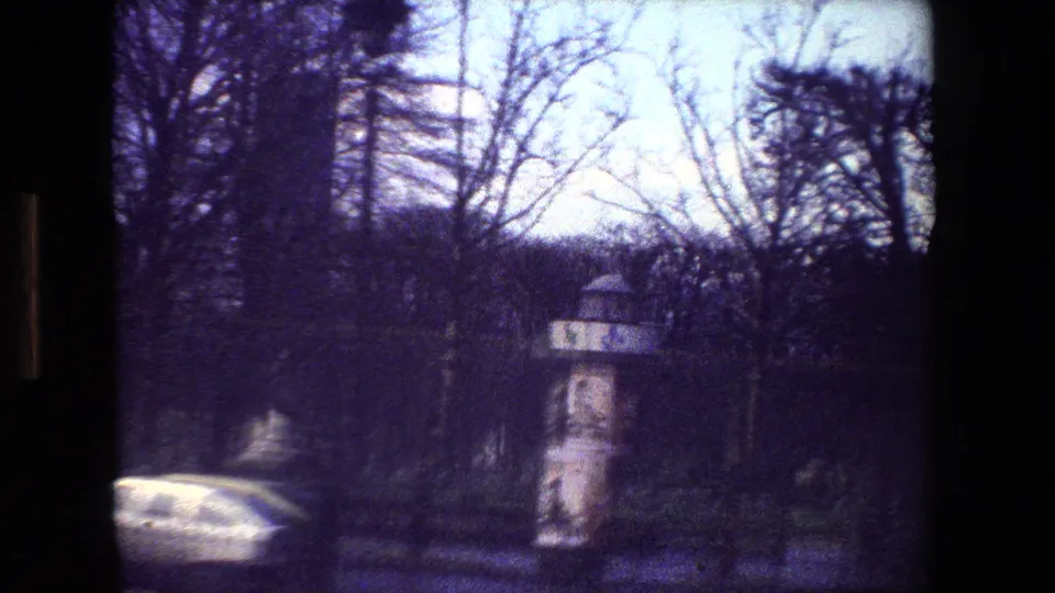 1981 Paris 8mm street view with a cylindrical advertising column or kiosk near the road, bare trees, a car, and soft handheld blur.