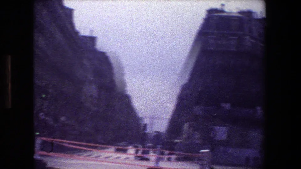 Paris city-street corridor in 1981 8mm footage, with tall buildings on both sides, road barriers, cars, and a cloudy pale sky.