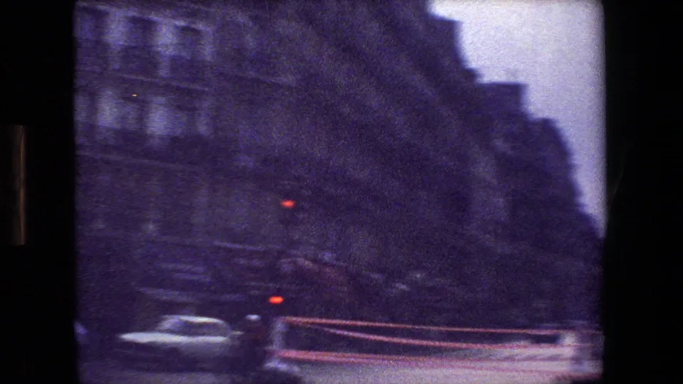 Early-1980s Paris street scene from a moving vehicle, with blurred cars at a red light, dark building facades, and visible 8mm frame edges.
