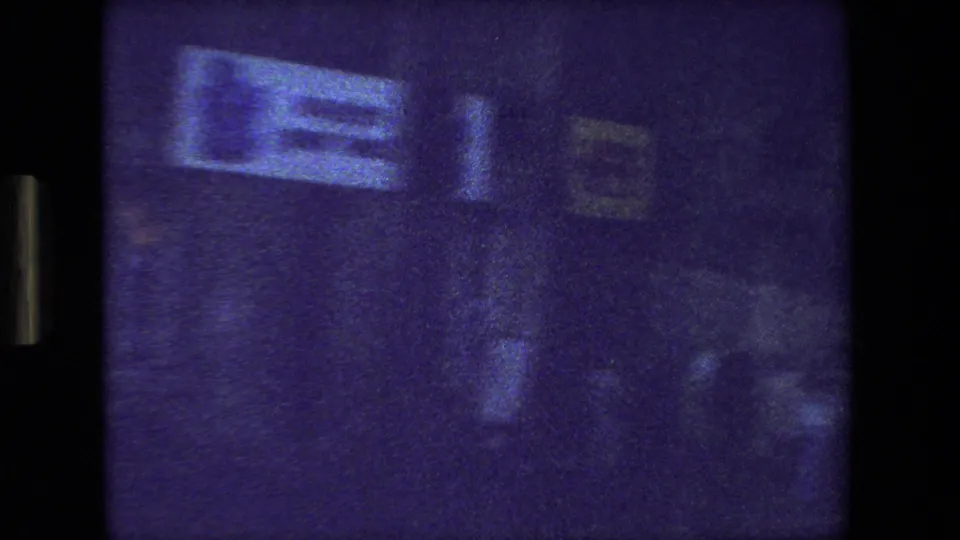 Grainy 1981 Paris frame with blurred reflections or street-side signage and indistinct human figures seen through a dark vehicle window.