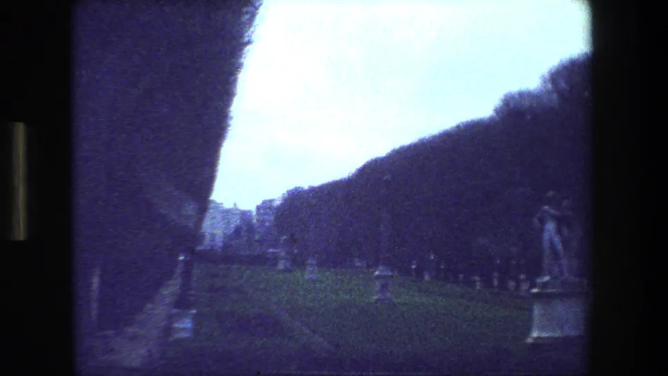 Moving 8mm view of a Paris park in 1981, with manicured grass, hedges, a path, distant buildings, and a statue near the right edge.