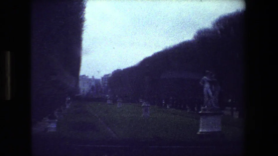1981 Paris 8mm frame showing a distant statue of a dancing couple in a cemetery, with grass, grave markers, and a dark tree-covered slope behind it.