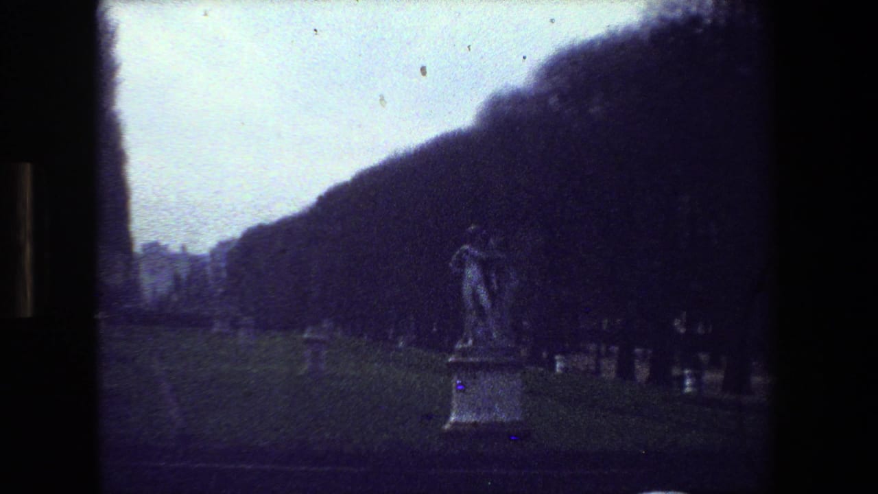 1981 Paris 8mm frame with the cemetery statue more centered, showing a dancing couple sculpture on a pedestal amid grass, graves, and dark trees.