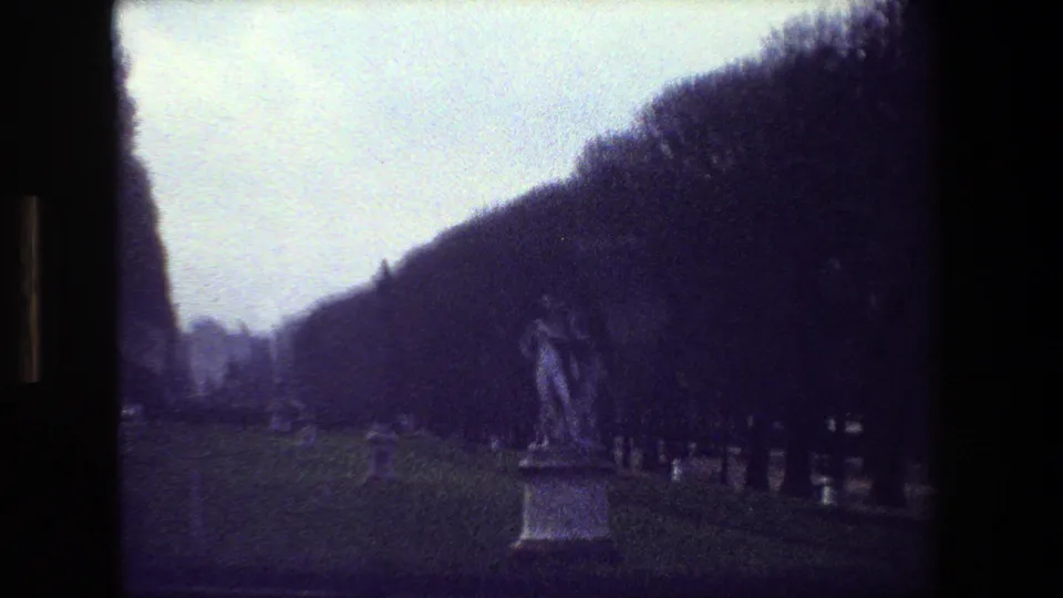 1981 Paris 8mm frame showing a closer visitor-shot view of the dancing couple cemetery statue, with grave markers along the path and dense dark trees in the background.