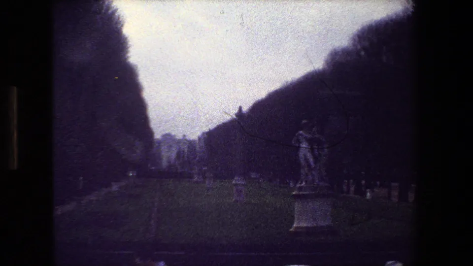 1981 Paris 8mm frame with the dancing couple statue positioned to the right, bordered by a grassy cemetery path, scattered grave markers, and a dark wooded backdrop.