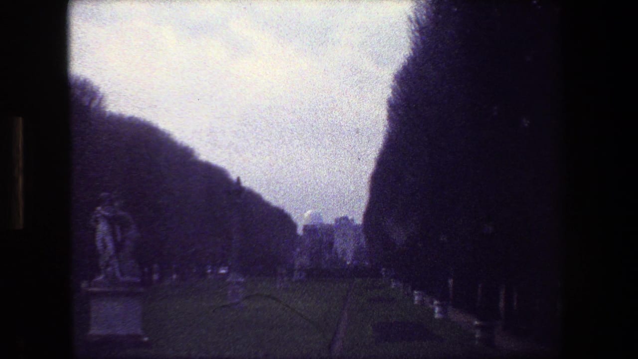 Paris, France 1981 home-movie frame showing a cemetery path bordered by dark trees, low hedges, classical statues, and grave markers under an overcast sky.