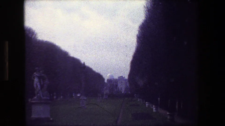 Paris, France 1981 home-movie frame showing a cemetery path bordered by dark trees, low hedges, classical statues, and grave markers under an overcast sky.
