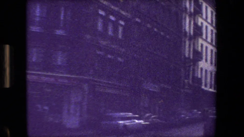 Paris, France 1981 home-movie frame shifting to a dim, blurred street view with tall building facades, reflective pavement, and strong purple color cast.
