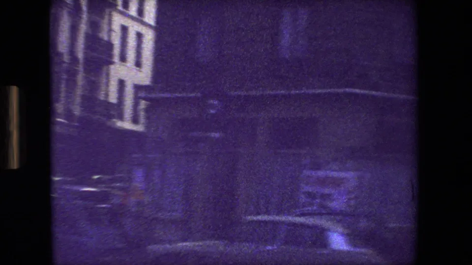 Paris, France 1981 frame showing an indistinct night street view with a building facade, dark pavement, bright reflections, and a parked car-like form.