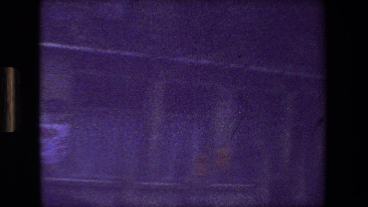 Purple-tinted 8mm frame from the Paris 1981 clip showing a blurred moving-vehicle view with dark borders, faint street reflections, and indistinct building surfaces.