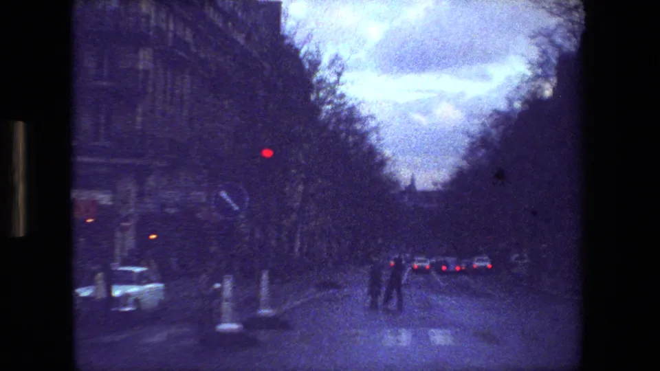 Paris home-movie frame from 1981 showing a broad boulevard with pedestrians in the roadway area, cars ahead, a red traffic light, and dark building facades.