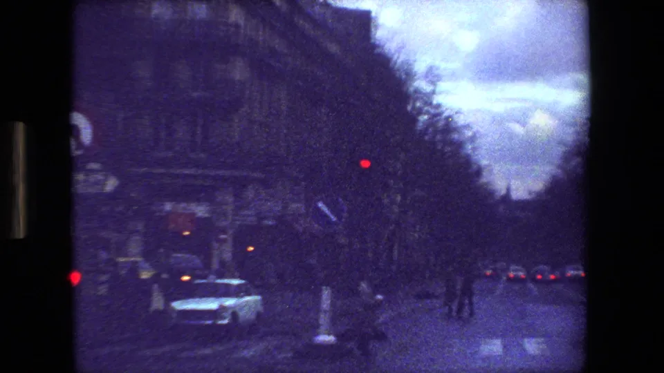 Paris street scene from the supplied 1981 8mm footage, showing a white car, red signal light, dark roadway, and rows of buildings at dusk.