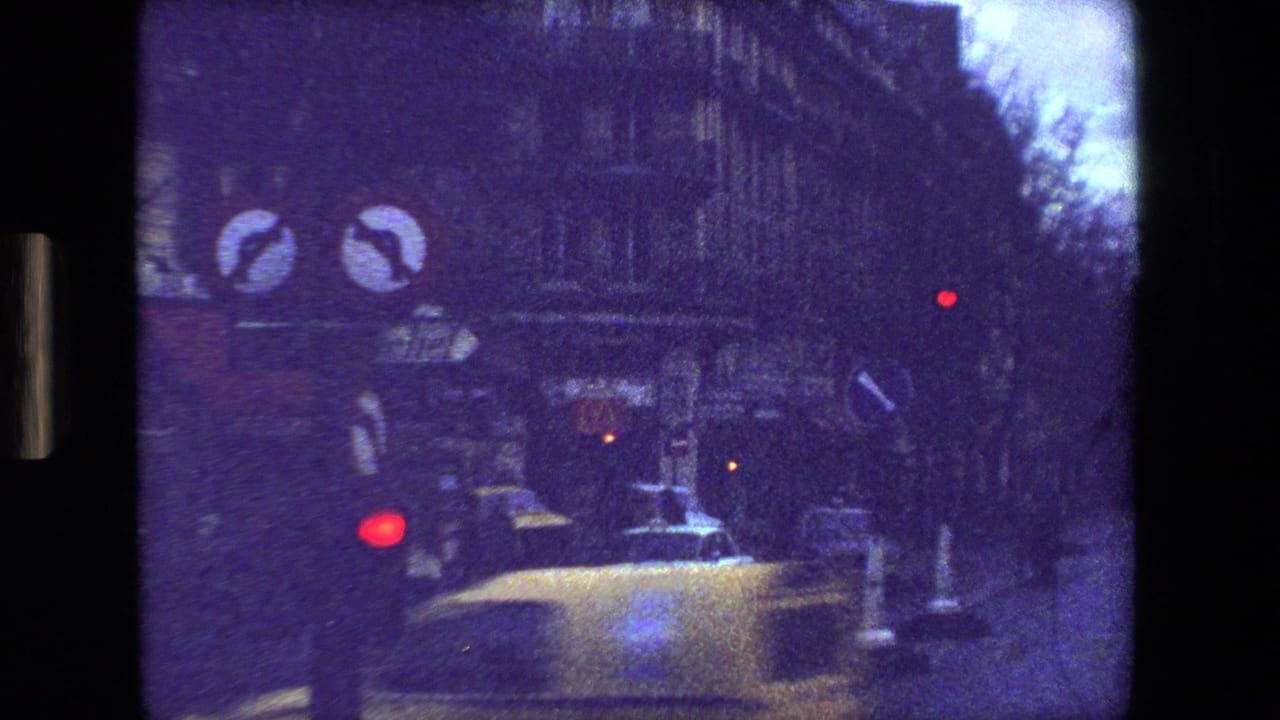 Forward-moving 1981 Paris street view with road signs, a light-colored car, red signal light, and dark building facades in grainy 8mm color.