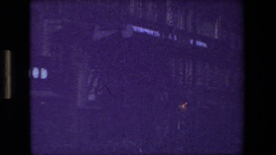 Underexposed frame from the Paris 8mm street sequence, with dark facades, roadway, faint distant lights, and the same blue-purple film cast.