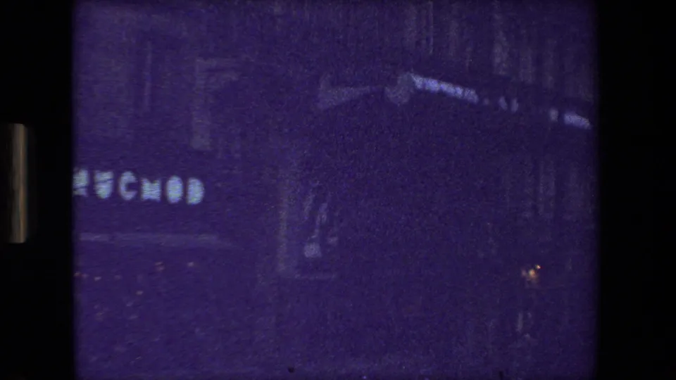 Grainy 1981 Paris street frame showing dark building fronts, partial street-level lettering, roadway shadows, and small points of traffic light.