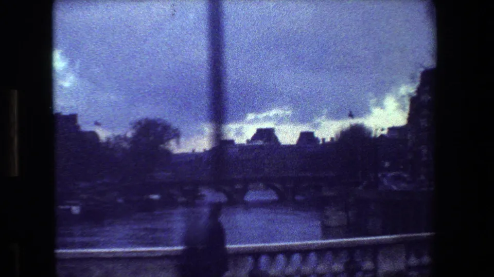 Grainy blue-tinted 1981 Paris riverside view with an arched bridge over the water, distant buildings, clouds, and a stone balustrade in the foreground.