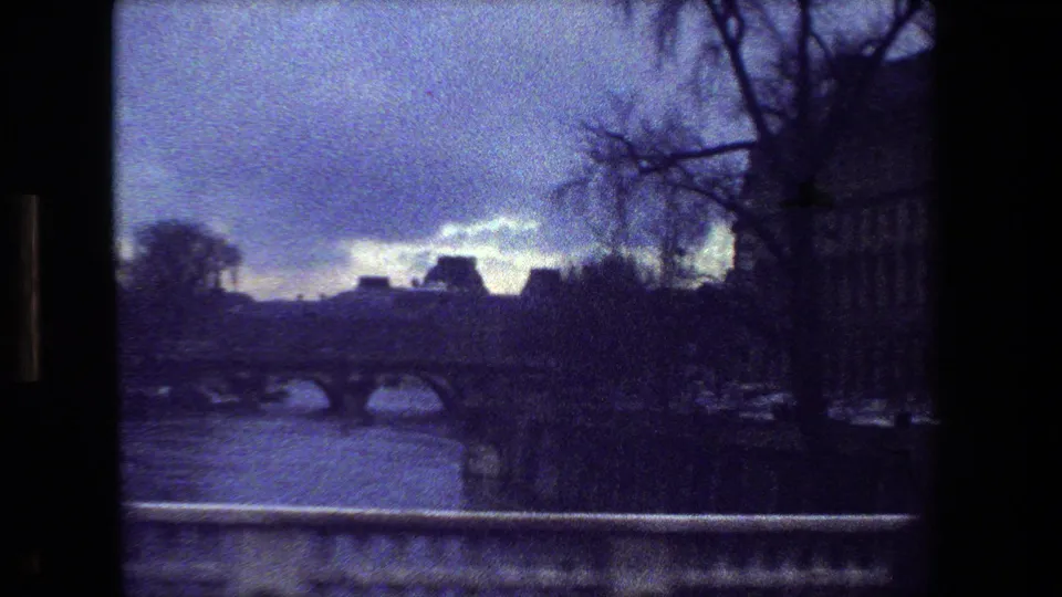 1981 Paris home-movie frame showing a river, low arched bridge, silhouetted leafless trees, distant roofline, and foreground stone railing in grainy 8mm color.
