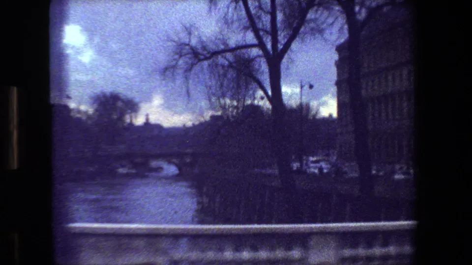 Dark blue 1981 Paris frame with a river or street-edge foreground, bare tree branches, building edge, stone railing, distant bridge area, and a small white car visible near the right side.
