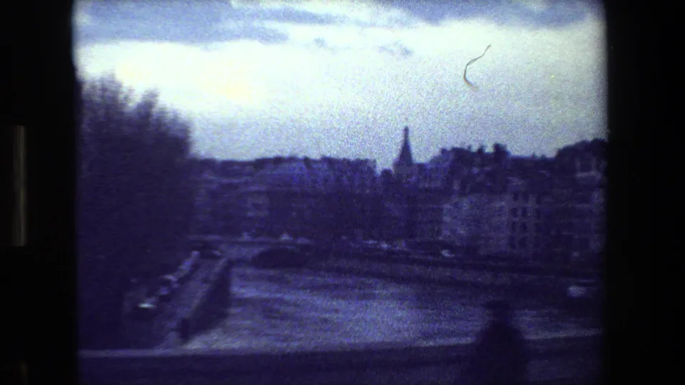 Blue-tinted Paris riverside view from the supplied 1981 home movie, showing a bridge edge, multi-story buildings, bare trees, and a distant spire under an overcast sky.