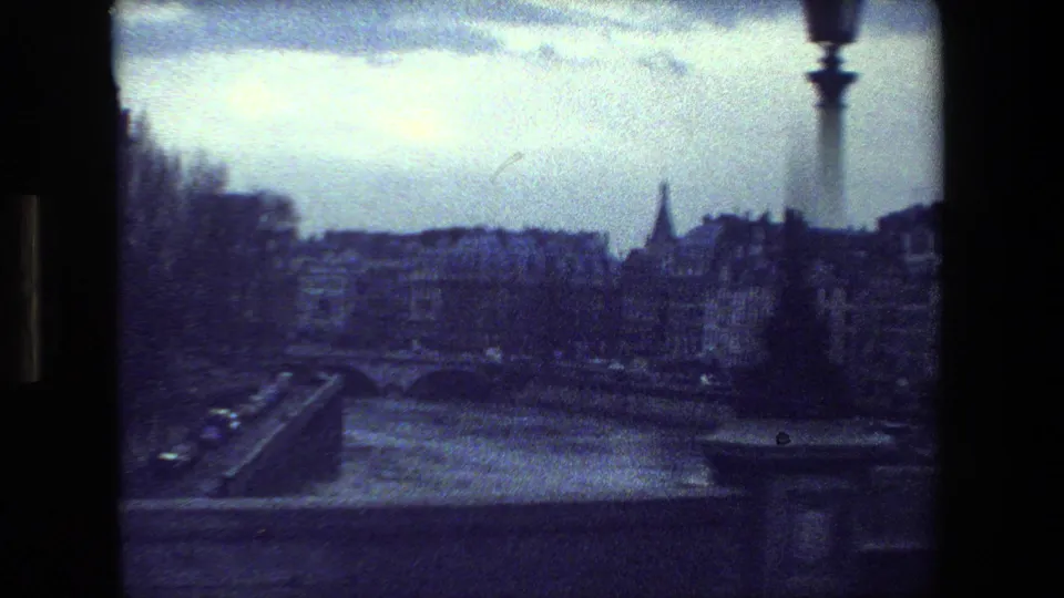 Moving-vehicle view in the supplied 1981 Paris 8mm footage, looking across a bridge and riverfront toward older buildings, a distant spire, and a vertical street fixture.