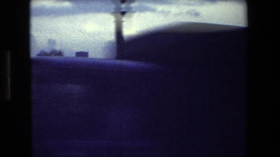 Obscured blue-tinted frame from the supplied Paris 1981 home movie, with a dark foreground edge, vertical pole, cloudy sky, and faint riverside structures in the distance.