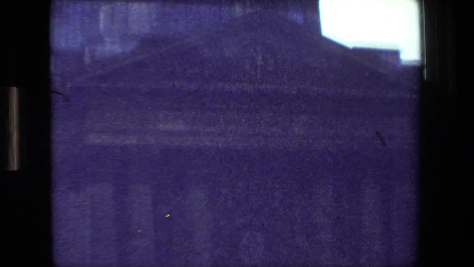 1981 Paris 8mm frame with shadowed riverfront buildings, a dark embankment or bridge area, grainy blue-purple exposure, and a bright patch of sky at the upper right.