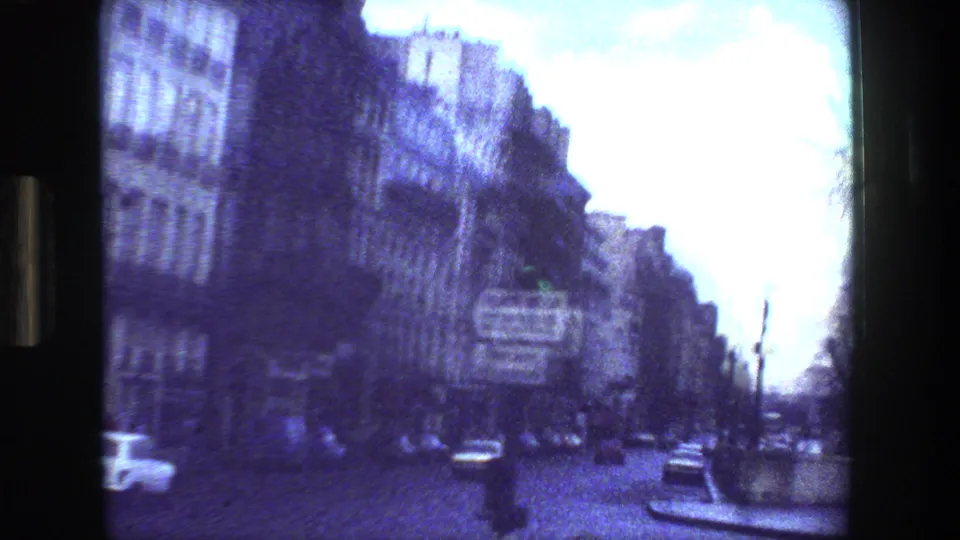 Grainy Paris street view from 1981 home-movie footage, with multi-story buildings, directional road signs, cars, and a bright overexposed sky.