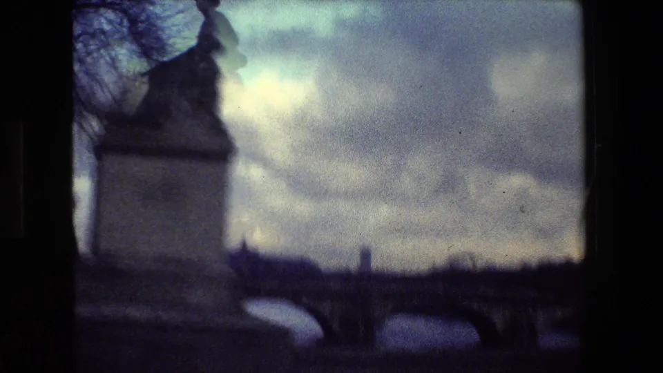 Soft 8mm Paris bridge view showing water, a stone bridge, cloudy sky, bare trees, and a dark statue or architectural silhouette in the foreground.