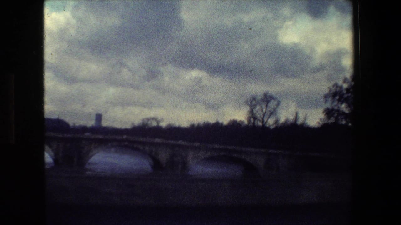 Paris, France 1981 home-movie frame showing a stone arch bridge crossing a river, with bare trees and a distant vertical structure under heavy clouds.