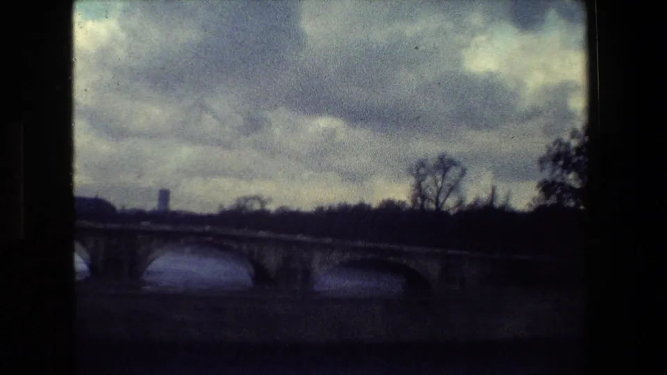Paris, France 1981 home-movie frame showing a stone arch bridge crossing a river, with bare trees and a distant vertical structure under heavy clouds.
