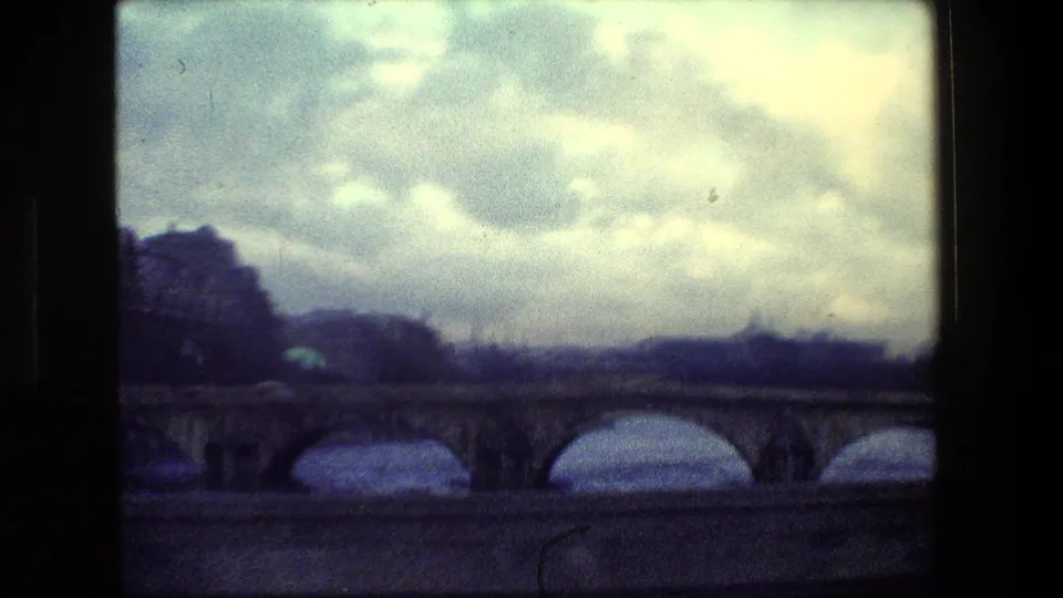 Paris, France 1981 home-movie frame showing a low, moving view past a stone arch bridge with river water below and a bright cloudy sky above.