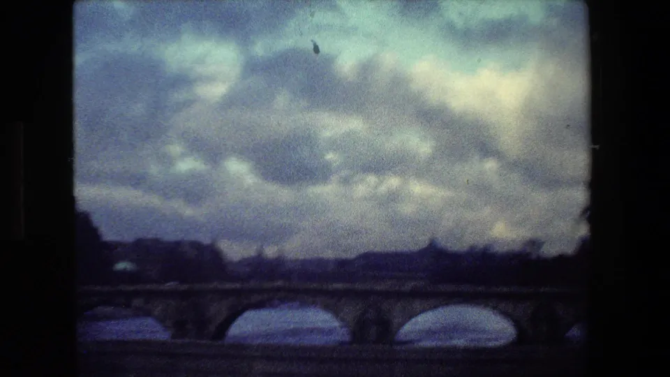 Paris, France 1981 frame showing a stone arch bridge over the river, with dark banks, distant low buildings, and dense cloud cover filling the upper frame.