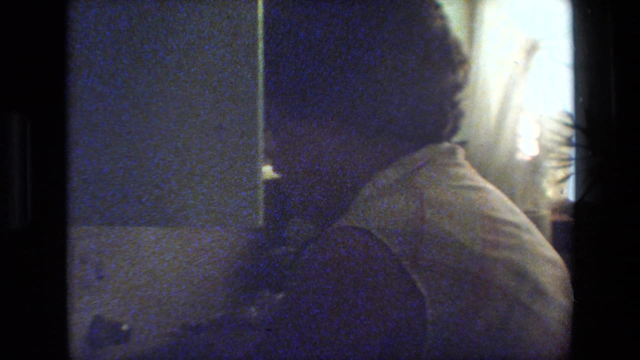 Grainy 8mm frame of an adult woman with glasses in a light sleeveless top, seen close to the camera in a dim private interior.