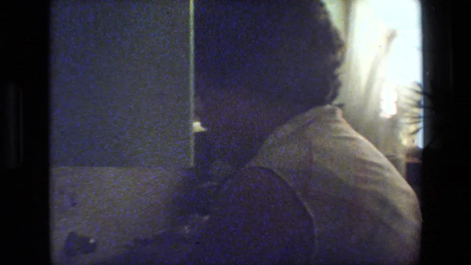 Grainy 8mm frame of an adult woman with glasses in a light sleeveless top, seen close to the camera in a dim private interior.