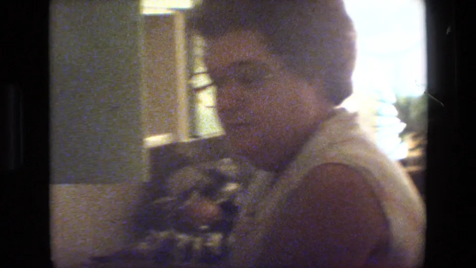 Soft 8mm frame of an adult woman with glasses facing toward the camera near household plants and interior surfaces.