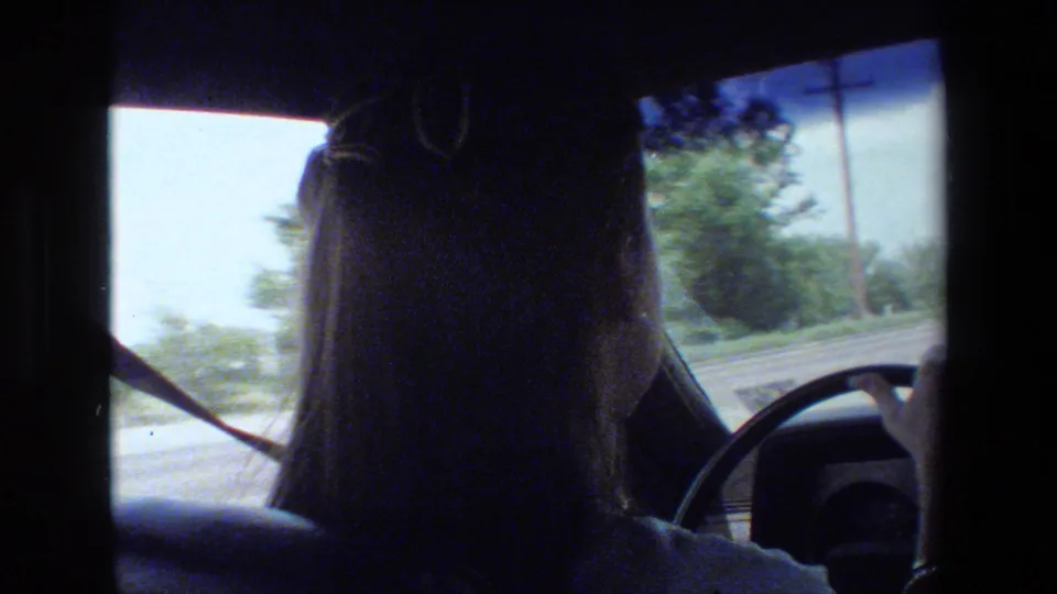 Home-movie view from inside a car showing the back of a driver at the steering wheel, with roadside trees and sky visible ahead.
