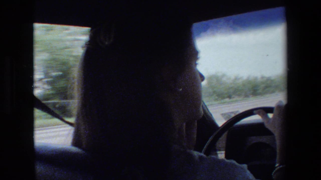 Young woman in side profile drives a car in archival 1981 home-movie footage, with the steering wheel, windshield, and roadside view visible.