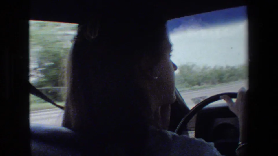 Young woman in side profile drives a car in archival 1981 home-movie footage, with the steering wheel, windshield, and roadside view visible.