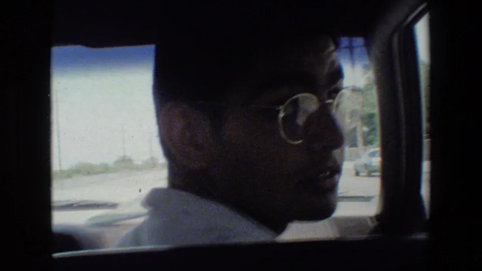 Young man wearing glasses looks toward the camera from the passenger area of a car, with window frames and a bright exterior view behind him.