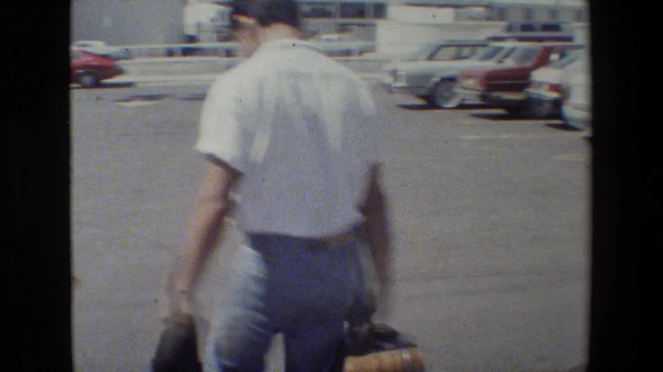 Man in a white shirt and blue jeans walks away through a parking lot carrying luggage, with parked cars visible in the background.