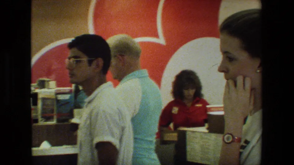 A young man in glasses and a white shirt waits near an airport counter while a red-uniformed woman works behind it, with a large red-and-white wall graphic in the background.
