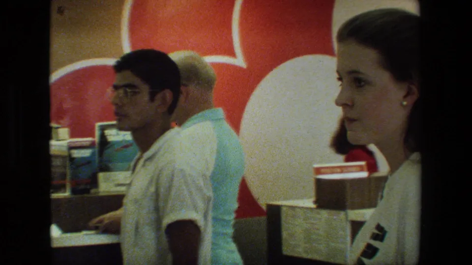 A woman in profile stands close to the camera at an airport counter area as a young man in glasses and another passenger wait near the service desk.