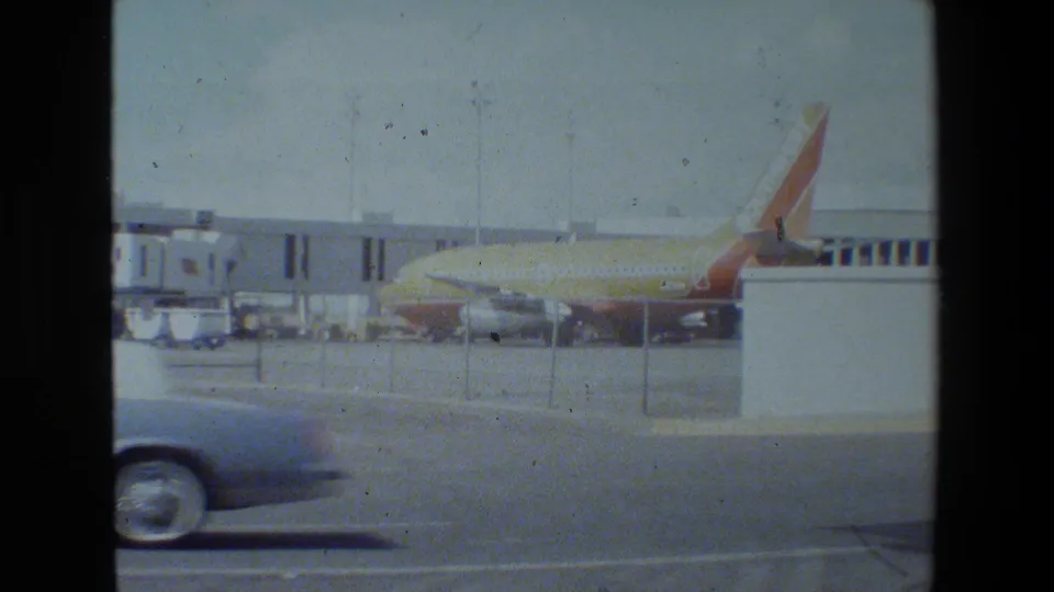 Through terminal windows, a parked commercial jet with a red and orange tail is visible beyond fencing while a car passes along the road in front.