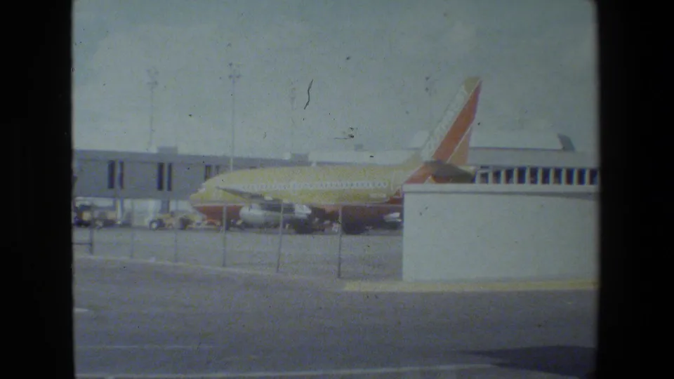 A commercial jet sits outside the terminal behind fencing, seen through dark window mullions and faded 8mm film grain.