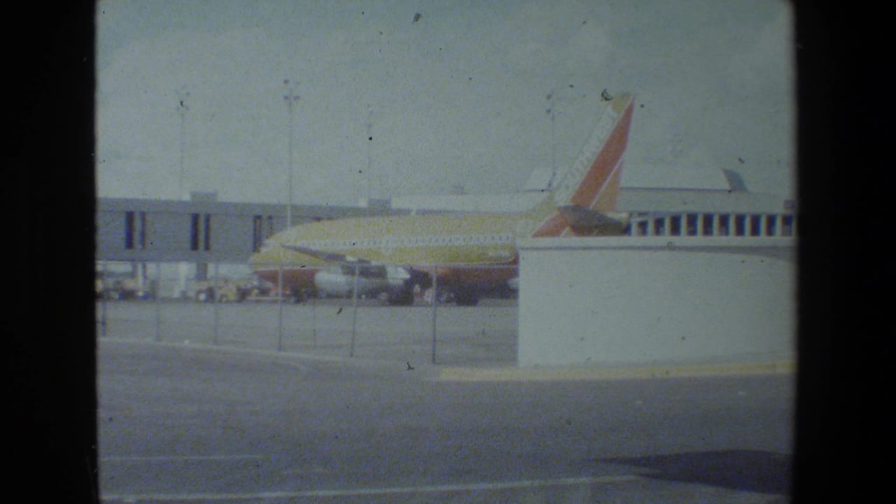 A Southwest-liveried passenger jet appears parked beside a terminal jet bridge on an airfield apron, filmed on grainy 8mm or Super 8 home-movie stock.