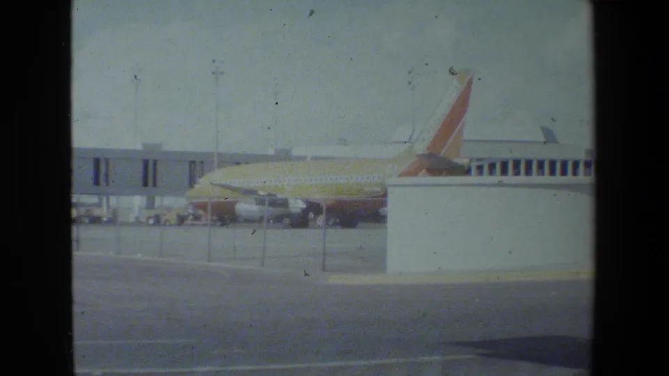 A Southwest-liveried passenger jet appears parked beside a terminal jet bridge on an airfield apron, filmed on grainy 8mm or Super 8 home-movie stock.