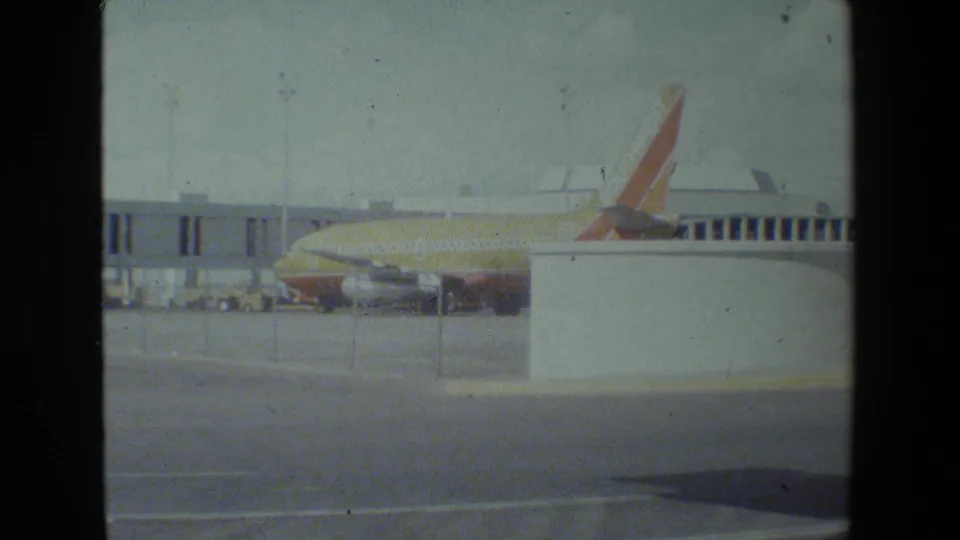 The aircraft remains parked at the terminal, with the red-orange tail visible above a white foreground structure and the jet bridge stretching behind it.