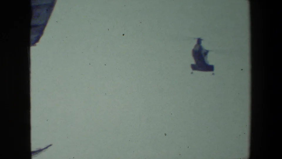 A helicopter crosses the pale sky near the airfield, with only a small portion of an aircraft wing or structure visible at the frame edge.