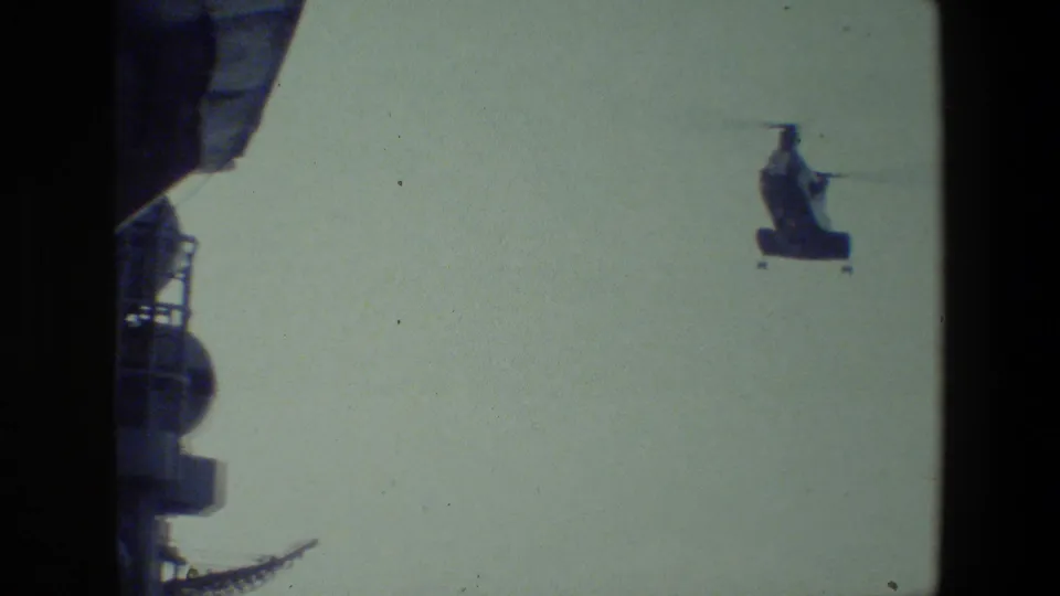 A distant tandem-rotor military helicopter appears in flight near the dark superstructure of a ship in metadata-supported 1981 8mm footage.